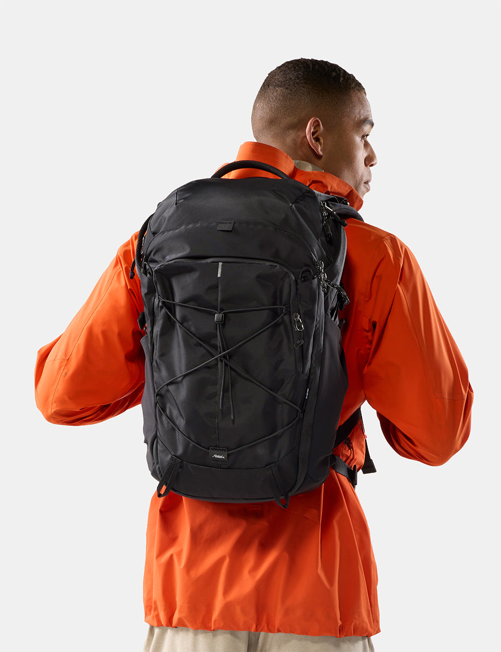 Matador 24L Adventure Pack in Black - a man wearing a bright orange jacket is shown from the back, carrying a black technical backpack with crisscross compression straps on a light gray background