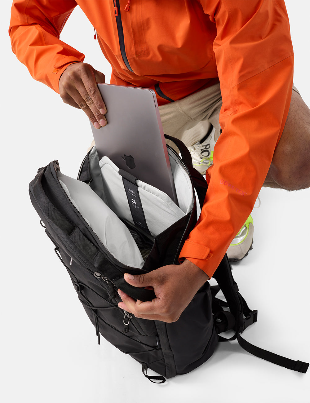 Matador 24L Adventure Pack in Black - person in orange jacket placing a laptop into a black backpack on a white background