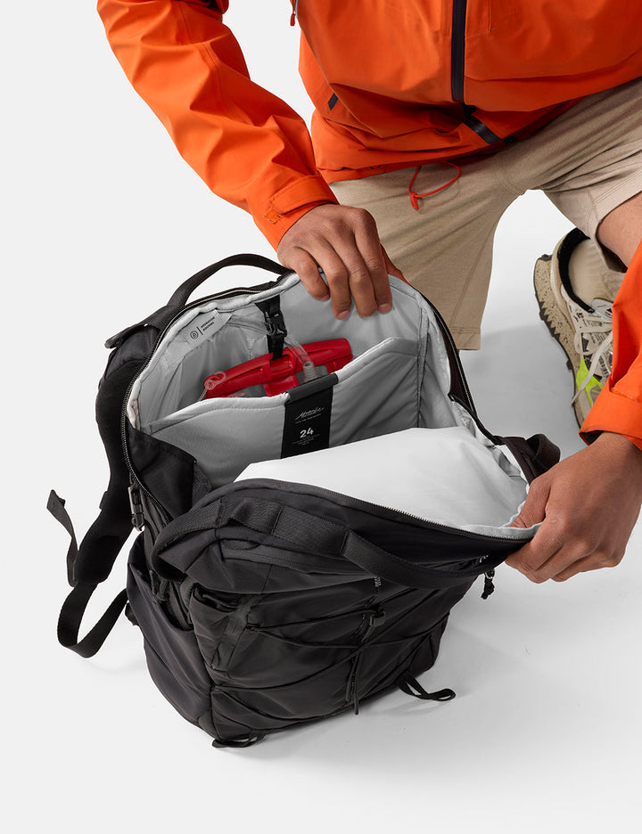 Matador 24L Adventure Pack in Black - person opening a black backpack with a white interior on a light gray background