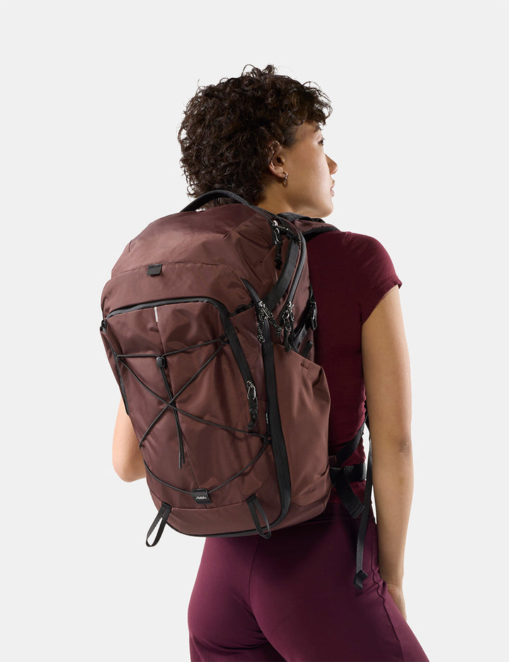 Matador 24L Adventure Pack in Garnet - a woman wearing a garnet hiking backpack with black straps, shown from a rear three-quarter view against a light gray studio background