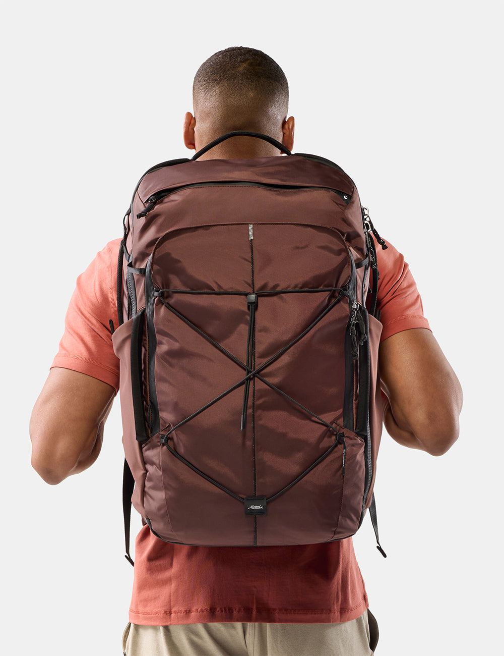 Matador 45L Global Travel Pack in Garnet - a man viewed from behind wearing a large garnet technical backpack with black compression straps and zippers, over a coral t-shirt, against a light gray background