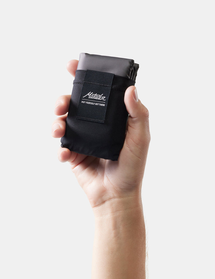 Matador Pocket Blanket™ 4.0 - 160 x 110cm in Black - hand holding a black pouch with a brand logo on a white background
