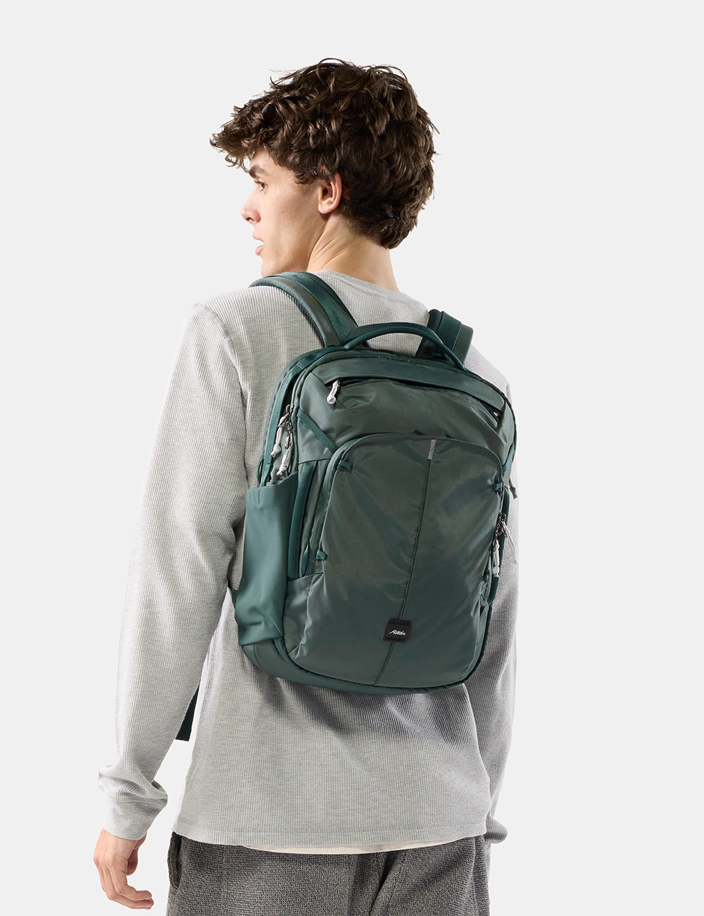 Matador 18L Personal Item Travel Pack in Juniper - a man wearing a gray sweatshirt and gray pants is shown from the back, wearing a dark green backpack. The photo is taken against a clean, light gray studio background