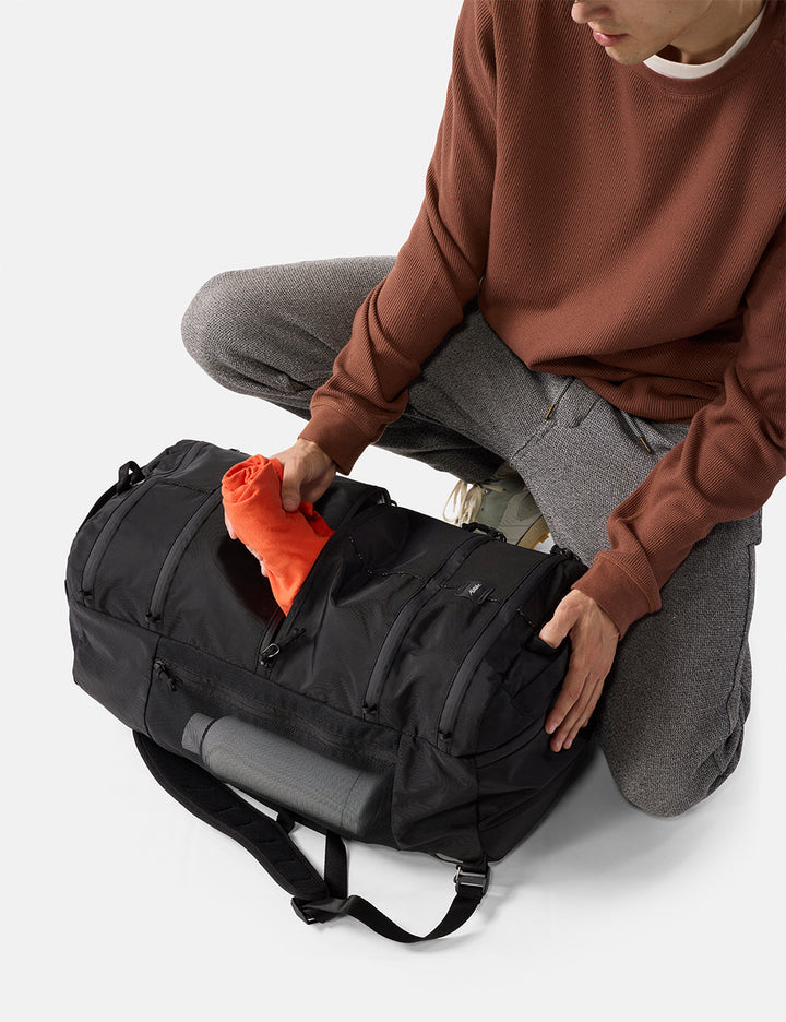 Matador 45L SEG Hybrid Travel Pack in Black - person packing a black duffel bag with an orange item on a light gray background