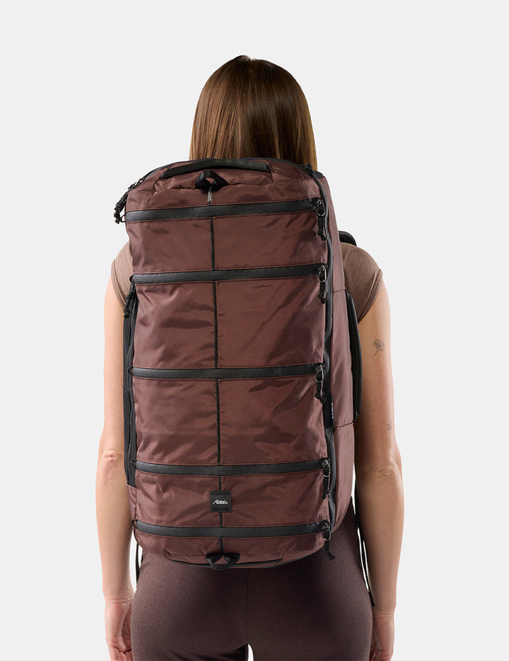 Matador 45L SEG Hybrid Travel Pack in Garnet - a woman viewed from behind wearing a large garnet segmented backpack against a light gray studio background