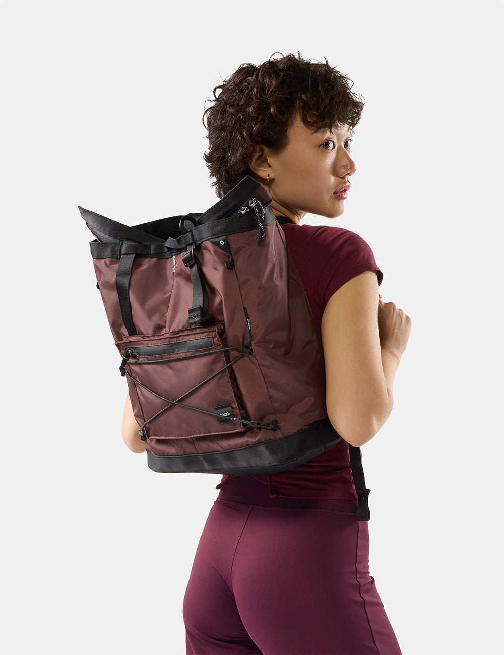 Matador 24L Tote Pack in Garnet - a woman wearing a garnet backpack with black trim and straps, shown from a side-back angle against a light gray studio background