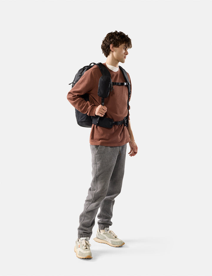 Matador Speed Stash (V2) in Black - person wearing a brown sweater, gray pants, and carrying a backpack on a white background
